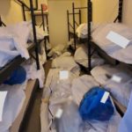 Body bags and blue bags containing the personal effects of the deceased are seen piled inside a portable refrigerator storage unit outside the hospital